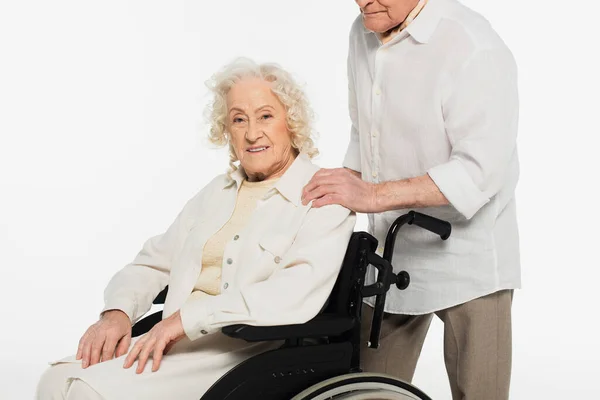 Sorridente donna anziana in sedia a rotelle vicino marito che tiene la mano sulla spalla isolata su bianco — Foto stock