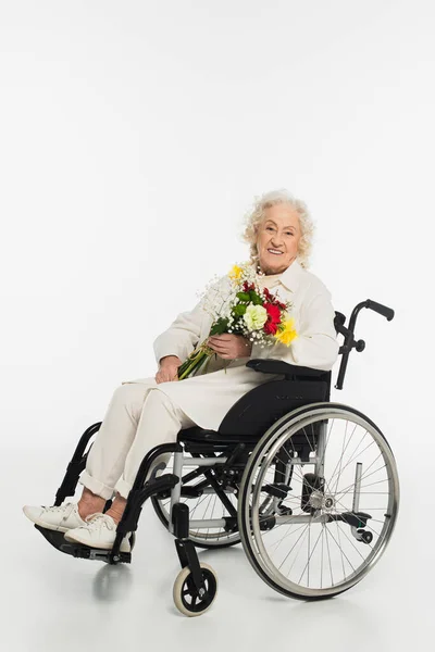 Smiling elderly woman in casual clothes sitting in wheelchair holding bouquet of flowers on white — Stock Photo
