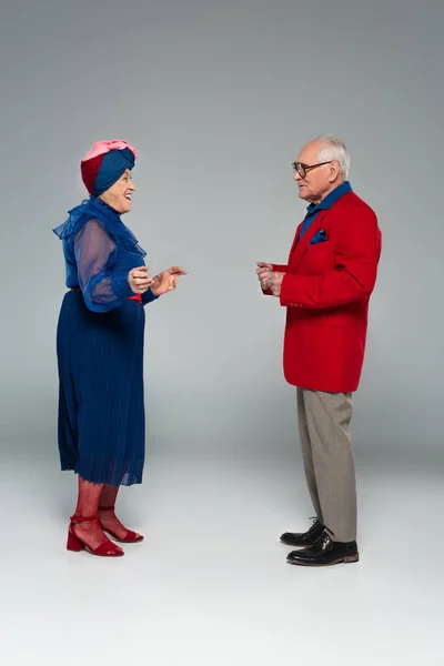 Улыбающаяся пожилая пара в синем платье и красный блейзер, танцующий на сером — стоковое фото
