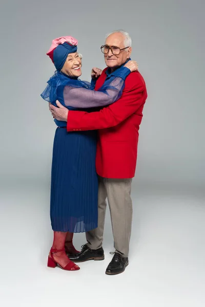 Sonriente pareja de ancianos en vestido azul y chaqueta roja abrazándose en gris - foto de stock