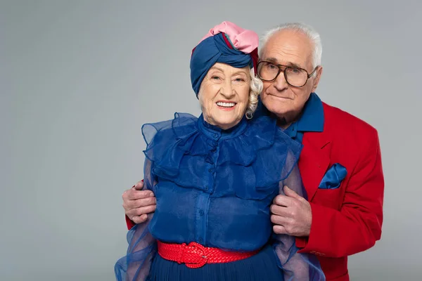 Sonriente anciano en blazer rojo abrazando esposa en vestido azul y turbante aislado en gris - foto de stock