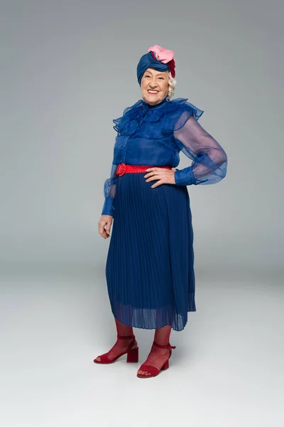 Smiling elderly woman in blue dress and turban posing with hand on hip on grey — Stock Photo