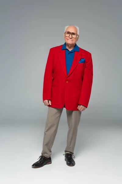 Full length view of serious elderly man in glasses, red blazer and blue shirt standing on grey — Stock Photo