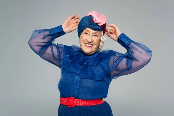 Femme âgée souriante en robe bleue ajustant turban avec les mains isolées sur gris — Photo de stock