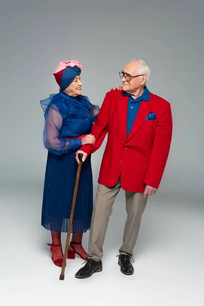 Smiling elderly woman in blue dress hugging husband in red blazer with walking stick on grey — Stock Photo