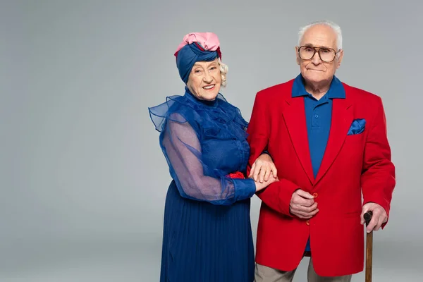 Lächelnde ältere Frau in blauem Kleid umarmt Ehemann im roten Blazer mit Spazierstock isoliert auf grau — Stockfoto