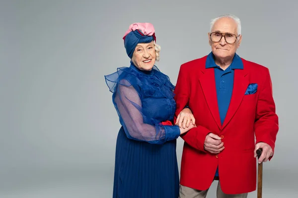 Sonriente anciana en vestido azul abrazando marido en blazer rojo con bastón aislado en gris - foto de stock