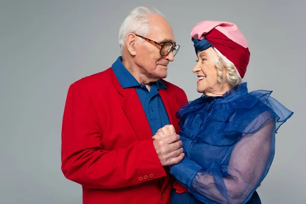 Sonriente anciano en blazer rojo cogido de la mano con la esposa en vestido azul y turbante aislado en gris - foto de stock