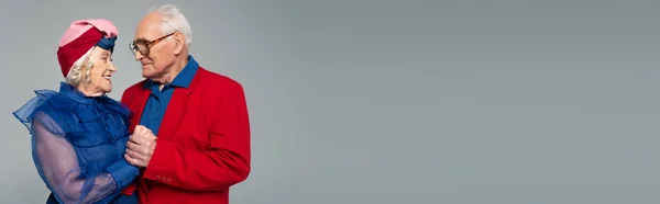 Sonriente anciano en blazer rojo cogido de la mano con la esposa en vestido azul y turbante aislado en gris, bandera - foto de stock