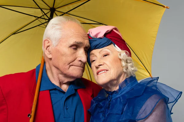 Homme âgé avec parapluie jaune étreignant femme en robe bleue et turban isolé sur gris — Photo de stock