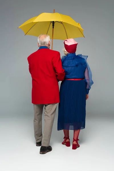 Back view of senior couple in stylish clothes walking with yellow umbrella on grey — Stock Photo