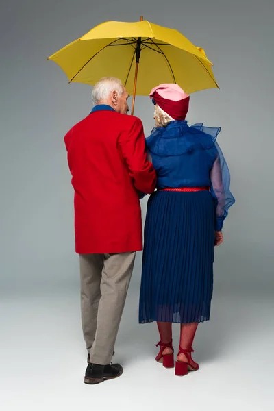 Back view of senior couple in stylish clothes walking with yellow umbrella on grey — Stock Photo