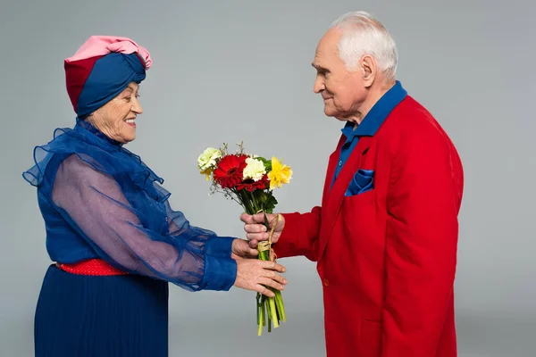 Smiling elderly man in red blazer presenting bouquet of flowers to wife in blue dress and turban isolated on grey — Stock Photo