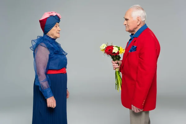Älterer Mann im roten Blazer hält Blumenstrauß neben Frau in blauem Kleid und Turban auf grau — Stockfoto