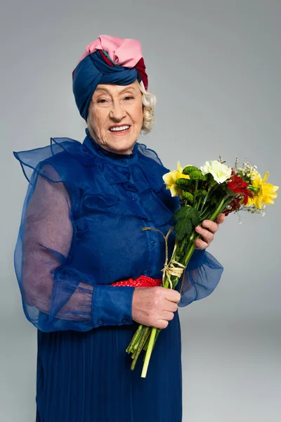 Smiling elderly woman in blue dress and turban holding bouquet of flowers isolated on grey — Stock Photo
