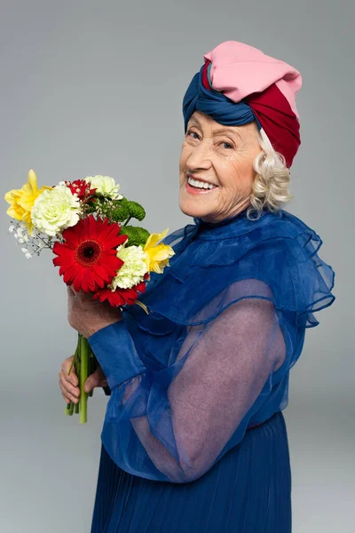 Mulher idosa positiva em vestido azul e turbante segurando buquê de flores isoladas em cinza — Stock Photo