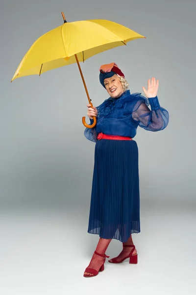 Smiling elderly woman in blue dress and turban holding yellow umbrella and waving with hand on grey — Stock Photo