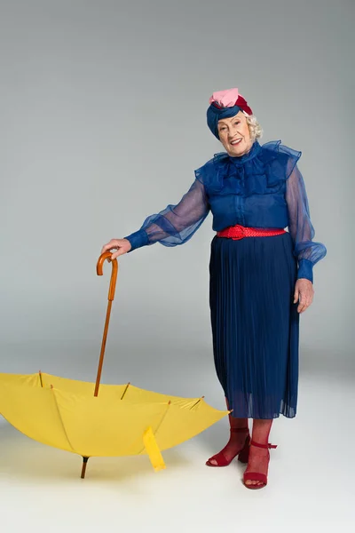 Sorrindo mulher idosa em vestido azul e turbante segurando guarda-chuva amarelo no cinza — Fotografia de Stock