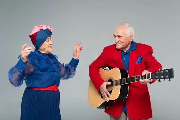 Smiling elderly man in red blazer playing acoustic guitar near dancing woman in blue dress isolated on grey — Stock Photo