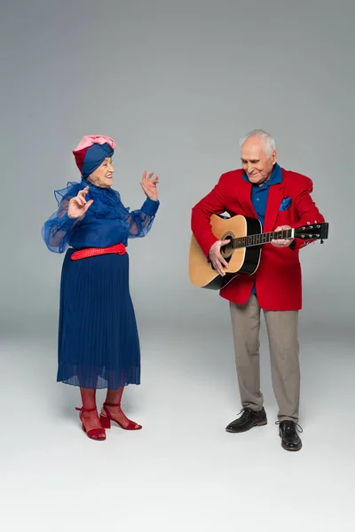 Hombre anciano positivo en chaqueta roja tocando la guitarra acústica cerca de la mujer bailarina en vestido azul y turbante en gris - foto de stock