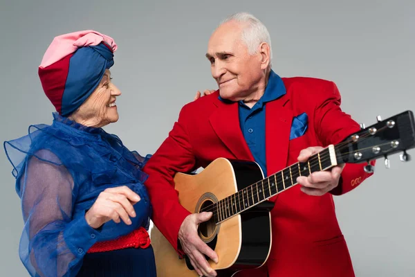 Sonriente anciano en chaqueta roja tocando la guitarra acústica cerca de la mujer bailarina en vestido azul y turbante aislado en gris - foto de stock