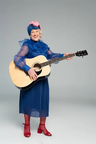 Sorrindo mulher idosa em vestido azul e turbante segurando guitarra acústica em cinza — Fotografia de Stock