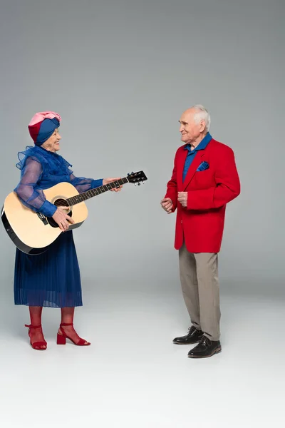 Positives älteres Paar in stylischer Kleidung tanzt mit Akustikgitarre auf grau — Stockfoto