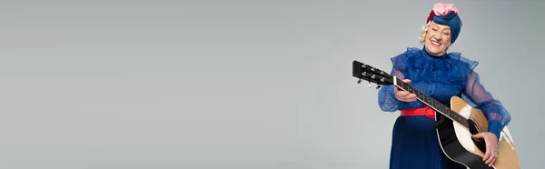 Усміхнена літня жінка в стильному одязі, що тримає акустичну гітару ізольовано на сірому, банер — стокове фото