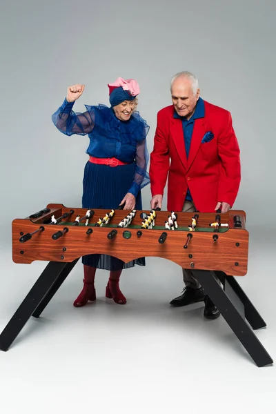 Pareja de ancianos emocionados en ropa elegante jugando al futbolín con gesto de victoria en gris - foto de stock