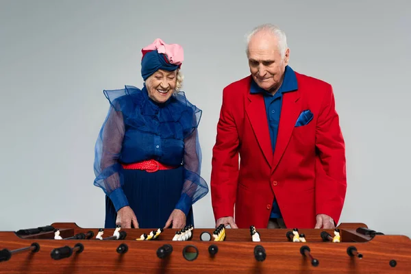 Positive elderly couple in stylish clothes playing table football on grey — Stock Photo
