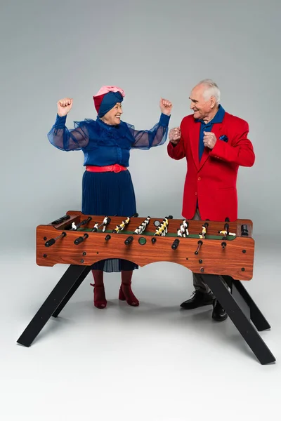 Excited elderly couple in stylish clothes playing table football with win gestures on grey — Stock Photo