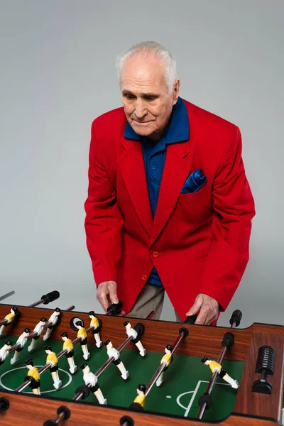 Senior man in red blazer playing table football on grey — Stock Photo