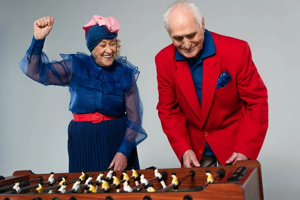 Excited elderly couple in stylish clothes playing table football with win gesture on grey — Stock Photo