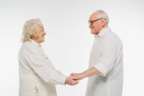 Sonriente pareja de ancianos en ropa casual tomados de la mano y mirándose aislados en blanco - foto de stock