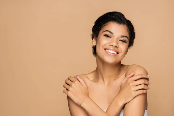 Mujer afroamericana feliz tocando hombros desnudos y mirando a la cámara aislada en beige - foto de stock