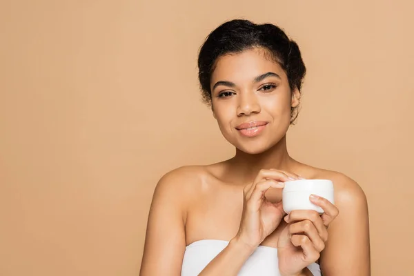 Femme afro-américaine souriante aux épaules nues tenant récipient avec crème cosmétique isolé sur beige — Photo de stock