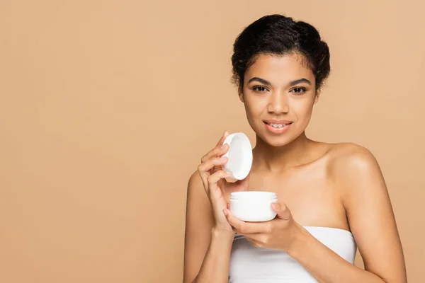 Joyful african american woman with bare shoulders holding container with cosmetic cream isolated on beige — Stock Photo