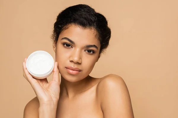 African american woman with bare shoulders holding container with face cream isolated on beige — Stock Photo