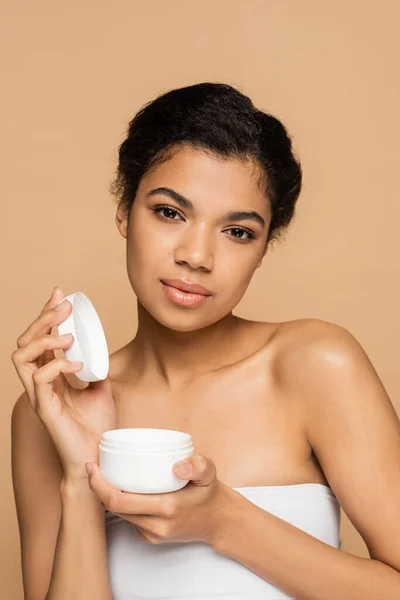 African american woman with bare shoulders holding container with face cream isolated on beige — Stock Photo