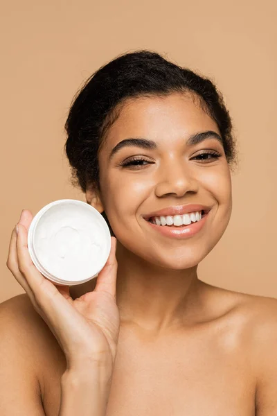 Pleased african american woman with bare shoulders holding container with face cream isolated on beige — Stock Photo