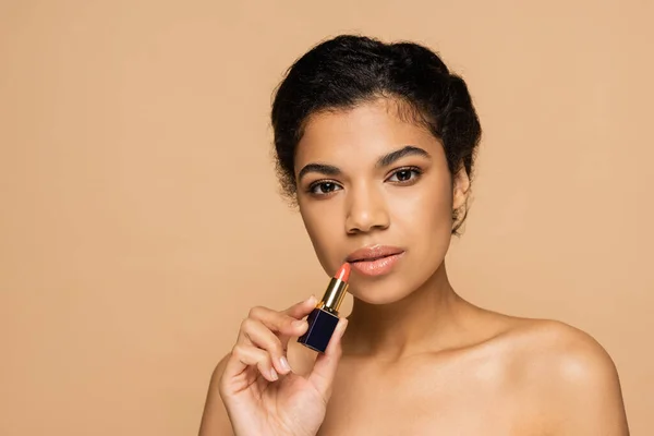 Mujer afroamericana con hombros desnudos sosteniendo lápiz labial aislado en beige - foto de stock