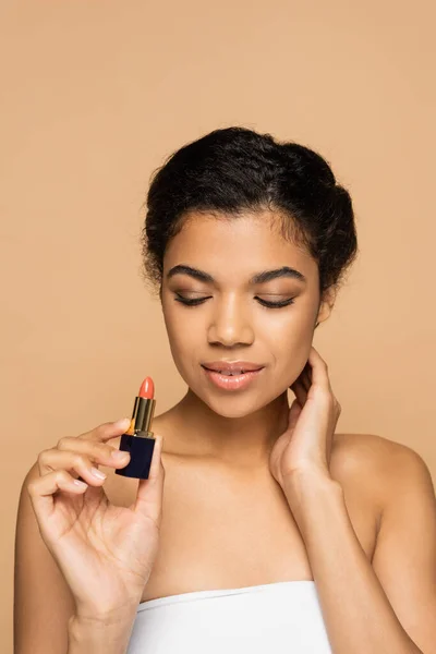 African american woman with bare shoulders looking at lipstick tube isolated on beige — Stock Photo