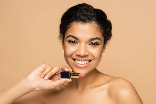 Joyful african american woman with bare shoulders holding lipstick isolated on beige — Stock Photo