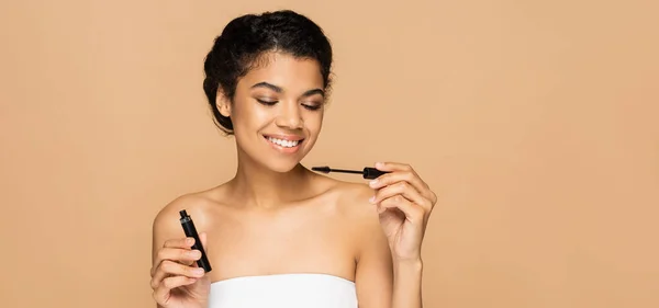 Happy african american woman with bare shoulders holding mascara tube and brush isolated on beige, banner — Stock Photo