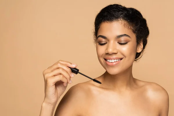 Femme afro-américaine souriante aux épaules nues tenant une brosse de mascara isolée sur beige — Photo de stock