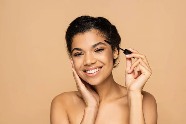 Cheerful african american woman with bare shoulders applying mascara isolated on beige — Stock Photo