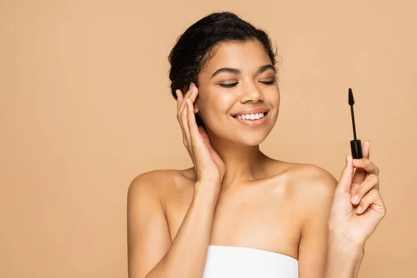 Joyeuse femme afro-américaine aux épaules nues tenant la brosse de mascara isolée sur beige — Photo de stock