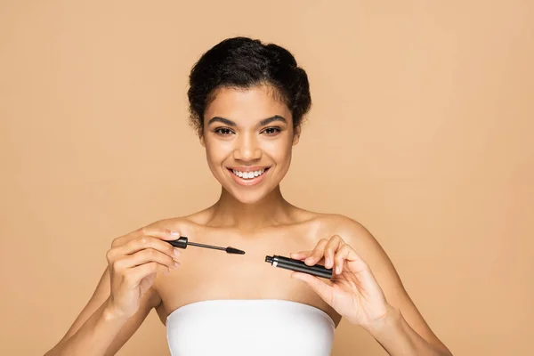 Positive femme afro-américaine aux épaules nues tenant tube de mascara et brosse isolée sur beige — Photo de stock