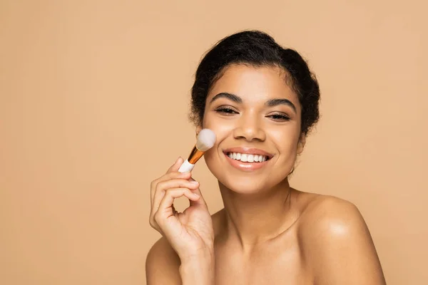 Glückliche afrikanisch-amerikanische Frau mit nackten Schultern, die Gesichtspuder mit Kosmetikbürste isoliert auf beige aufträgt — Stockfoto