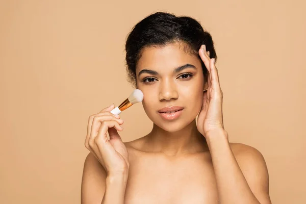 Junge afrikanisch-amerikanische Frau mit nackten Schultern, die Gesichtspuder mit Kosmetikbürste isoliert auf beige aufträgt — Stockfoto